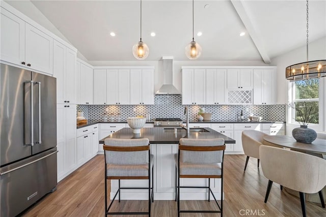 kitchen with pendant lighting, wall chimney range hood, high quality fridge, and an island with sink