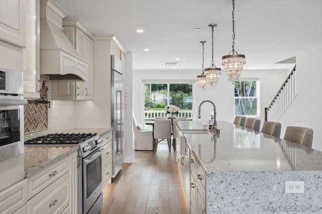 kitchen featuring an island with sink, appliances with stainless steel finishes, premium range hood, and pendant lighting