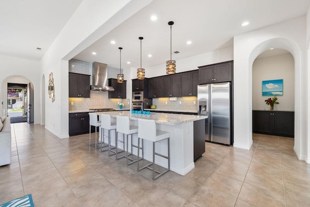 kitchen featuring backsplash, wall chimney range hood, dark brown cabinetry, stainless steel refrigerator with ice dispenser, and a center island with sink