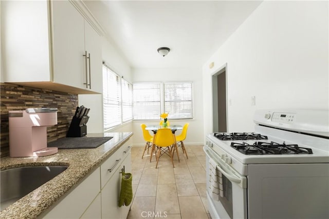 kitchen featuring light tile patterned flooring, white range with gas stovetop, white cabinets, light stone countertops, and backsplash