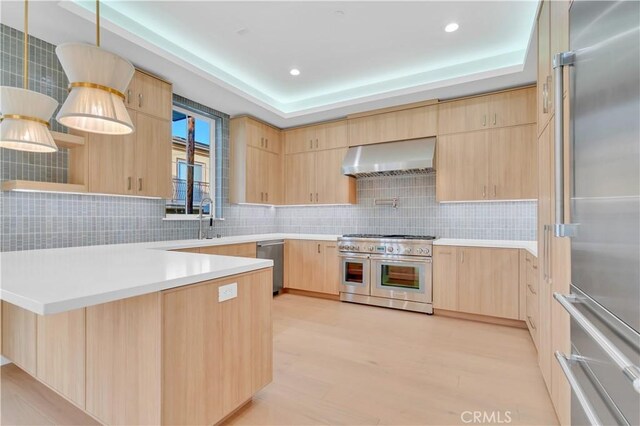 kitchen featuring pendant lighting, high quality appliances, a tray ceiling, wall chimney exhaust hood, and light brown cabinets