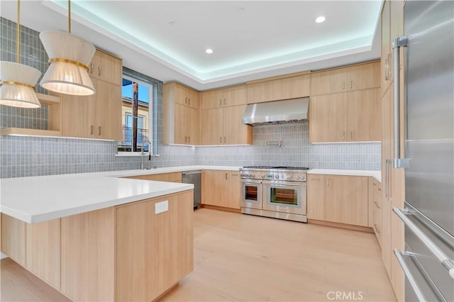 kitchen with pendant lighting, premium appliances, a tray ceiling, light brown cabinets, and wall chimney exhaust hood