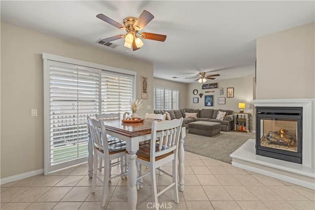 dining area with light tile patterned floors, a multi sided fireplace, visible vents, a ceiling fan, and baseboards