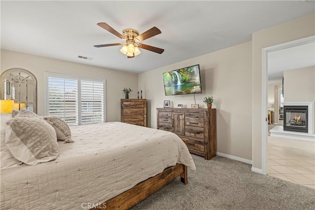 bedroom with light colored carpet, visible vents, a ceiling fan, a multi sided fireplace, and baseboards