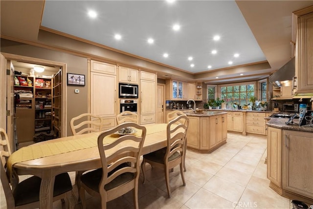 kitchen featuring a raised ceiling, wall chimney range hood, a kitchen island with sink, stainless steel appliances, and light brown cabinets