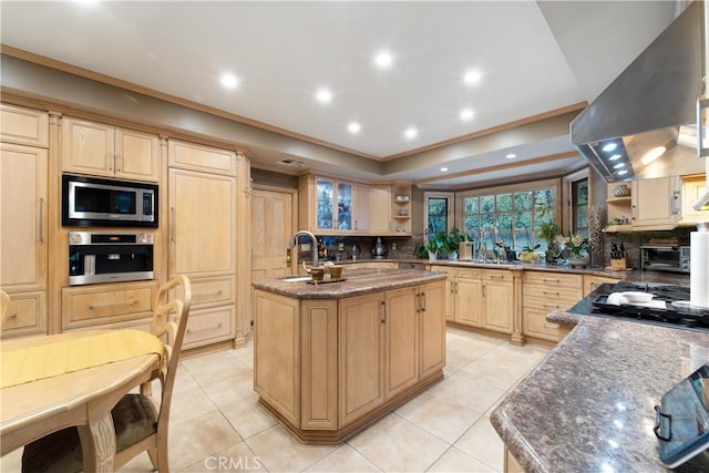 kitchen featuring a center island with sink, dark stone counters, light brown cabinets, stainless steel appliances, and range hood