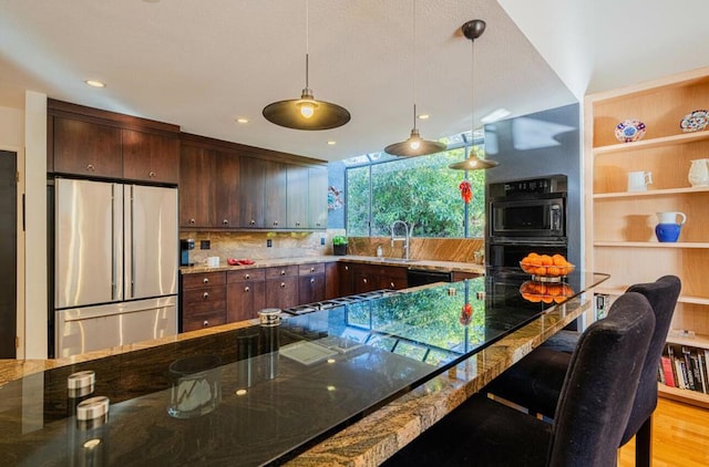 kitchen with sink, stainless steel refrigerator, stone counters, hanging light fixtures, and dark brown cabinetry