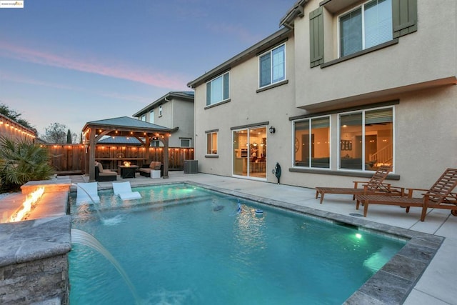 pool at dusk with a gazebo, pool water feature, an outdoor fire pit, and a patio