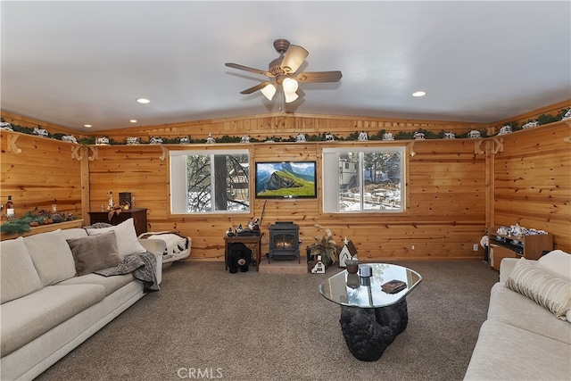 carpeted living room with vaulted ceiling, wood walls, ceiling fan, and a wood stove