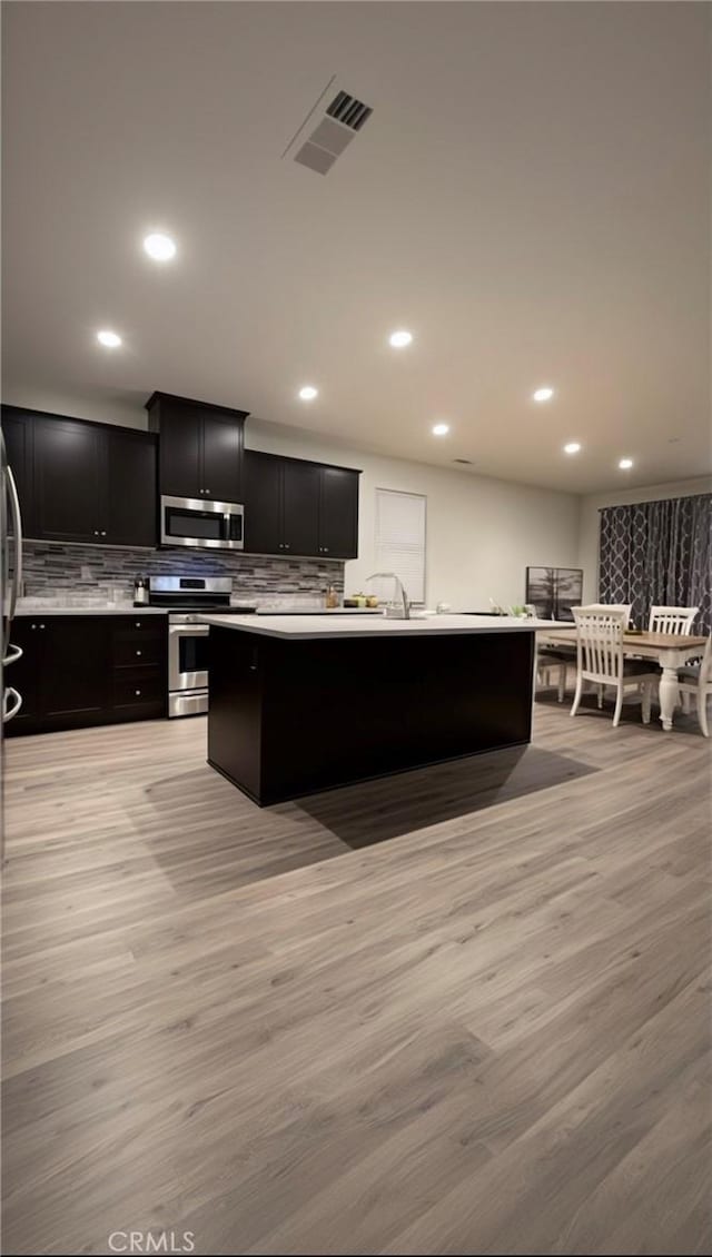 kitchen with light wood-type flooring, backsplash, a center island with sink, and appliances with stainless steel finishes