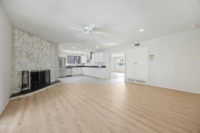 unfurnished living room with ceiling fan, a stone fireplace, a textured ceiling, and light wood-type flooring