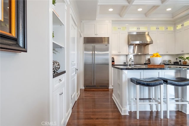 kitchen with a kitchen bar, built in refrigerator, beam ceiling, range hood, and white cabinets