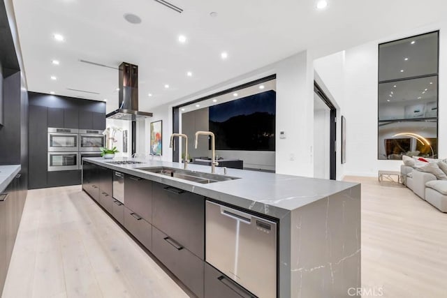 kitchen with sink, light hardwood / wood-style flooring, appliances with stainless steel finishes, island range hood, and a large island with sink