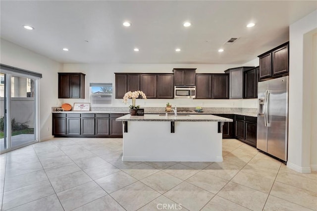 kitchen featuring a breakfast bar, light stone counters, stainless steel appliances, dark brown cabinets, and a center island with sink