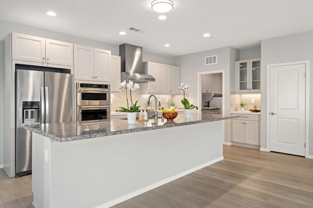 kitchen with a kitchen island with sink, wall chimney range hood, stainless steel appliances, and white cabinets