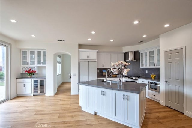 kitchen with wine cooler, white cabinetry, a center island with sink, light hardwood / wood-style floors, and wall chimney range hood