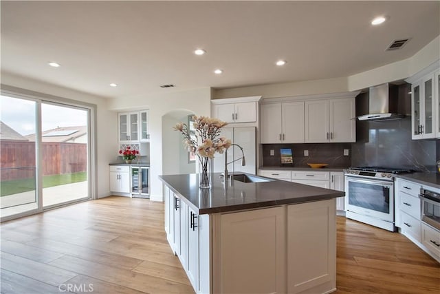 kitchen with wall chimney range hood, sink, stainless steel appliances, an island with sink, and white cabinets