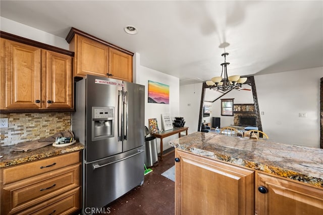 kitchen featuring brown cabinets, tasteful backsplash, an inviting chandelier, a stone fireplace, and stainless steel fridge