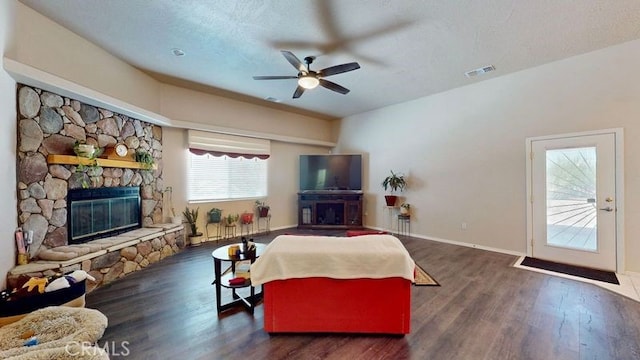 living room featuring ceiling fan, a stone fireplace, dark wood-type flooring, and a textured ceiling