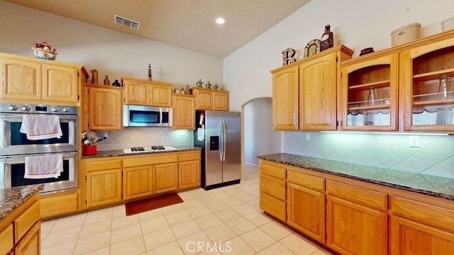 kitchen with appliances with stainless steel finishes, lofted ceiling, backsplash, dark stone counters, and light tile patterned floors