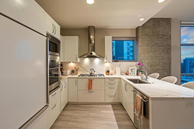 kitchen featuring appliances with stainless steel finishes, sink, light hardwood / wood-style floors, kitchen peninsula, and wall chimney range hood