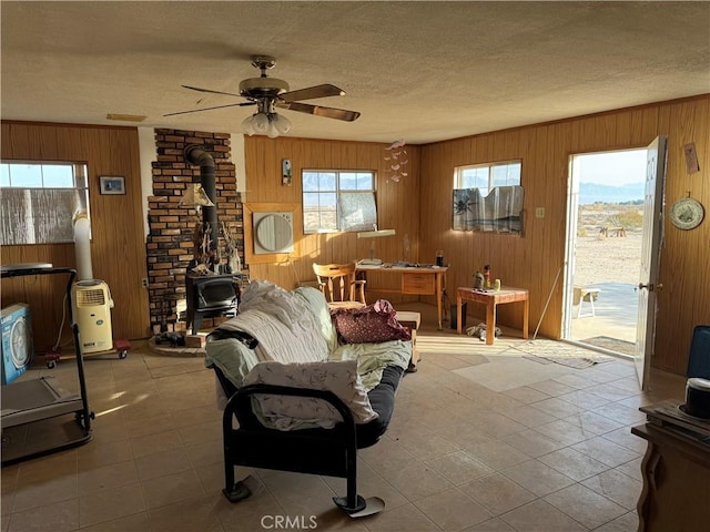 living room with ceiling fan, wooden walls, a textured ceiling, and a wood stove