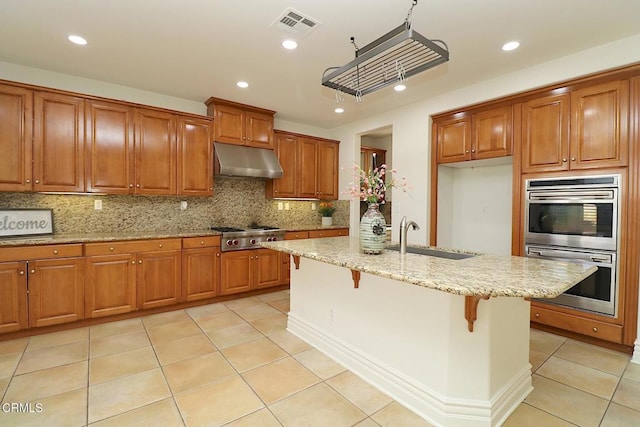 kitchen featuring sink, a breakfast bar, appliances with stainless steel finishes, light stone countertops, and an island with sink