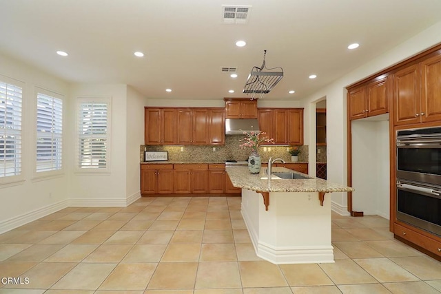 kitchen featuring double oven, an island with sink, sink, a breakfast bar area, and light stone countertops