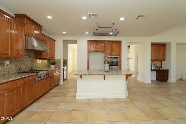 kitchen with a breakfast bar, a kitchen island with sink, backsplash, light stone counters, and stainless steel gas stovetop