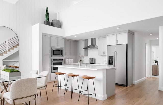 kitchen with light wood-type flooring, stainless steel appliances, wall chimney exhaust hood, and a center island with sink