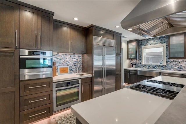 kitchen with appliances with stainless steel finishes, sink, decorative backsplash, island exhaust hood, and dark brown cabinets