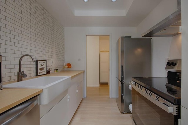 kitchen featuring appliances with stainless steel finishes, sink, a raised ceiling, light wood-type flooring, and wall chimney exhaust hood