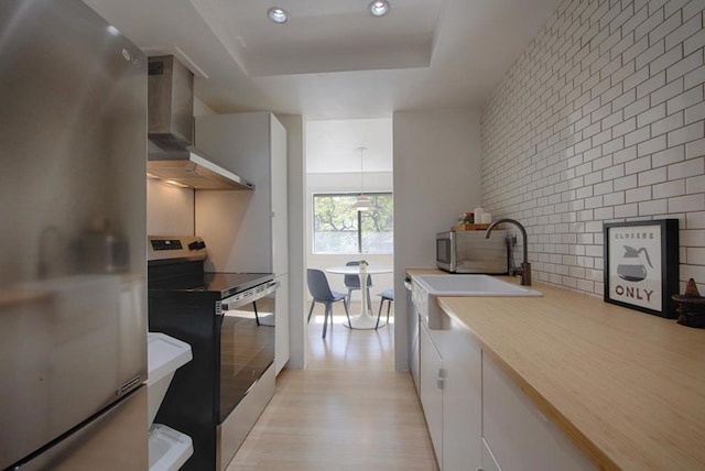 kitchen featuring a raised ceiling, white cabinets, wall chimney exhaust hood, stainless steel appliances, and light hardwood / wood-style flooring