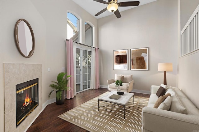 living room with dark wood-type flooring, ceiling fan, a fireplace, and a towering ceiling