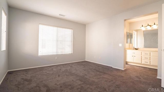 unfurnished bedroom featuring multiple windows, dark colored carpet, and ensuite bathroom