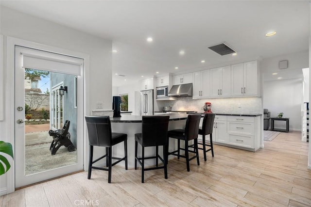 kitchen featuring dark countertops, appliances with stainless steel finishes, under cabinet range hood, a kitchen bar, and white cabinetry