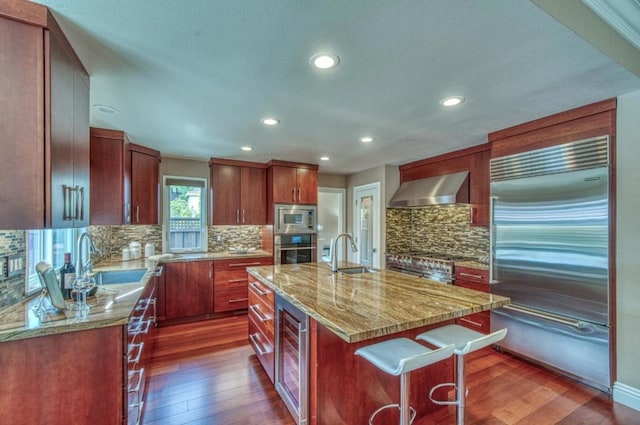 kitchen featuring built in appliances, sink, a center island with sink, and wall chimney range hood