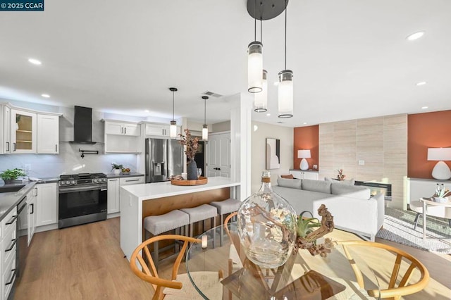 kitchen with hanging light fixtures, appliances with stainless steel finishes, white cabinets, wall chimney range hood, and backsplash