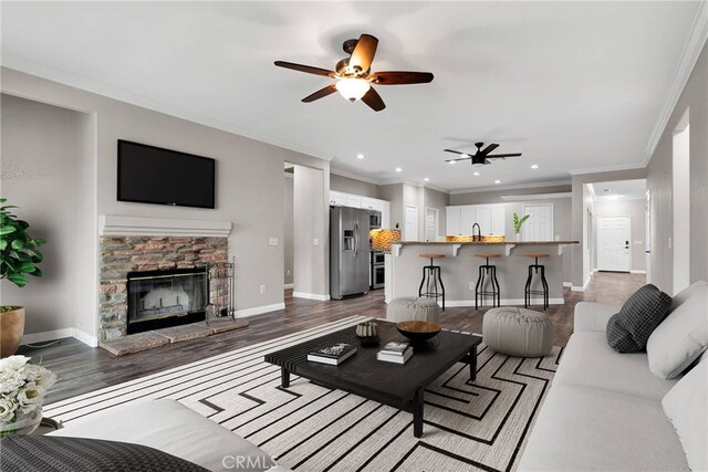 living room with a stone fireplace, sink, ceiling fan, crown molding, and dark wood-type flooring