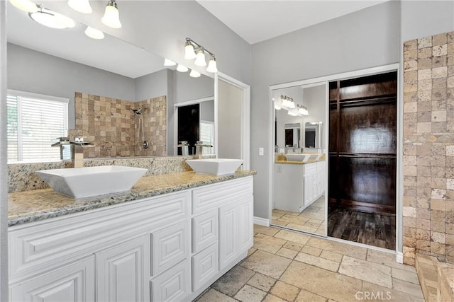full bath with a tile shower, stone tile floors, a sink, and double vanity