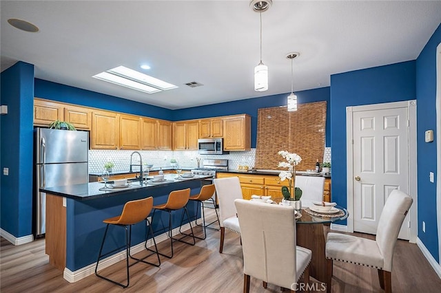 kitchen featuring appliances with stainless steel finishes, decorative light fixtures, decorative backsplash, a kitchen island with sink, and light wood-type flooring