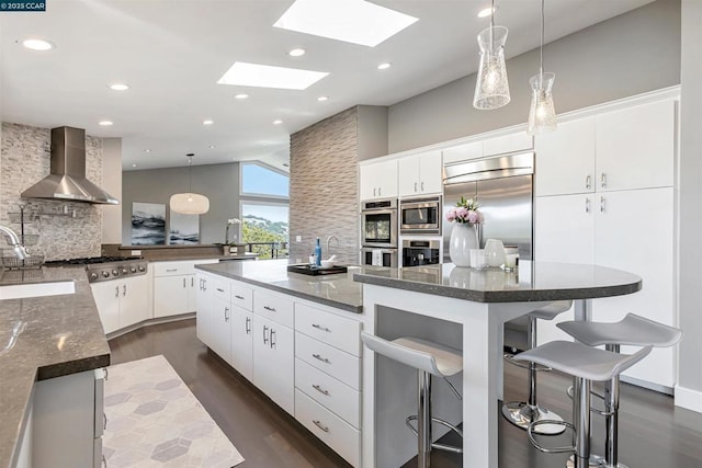 kitchen featuring a breakfast bar area, backsplash, white cabinets, built in appliances, and wall chimney range hood