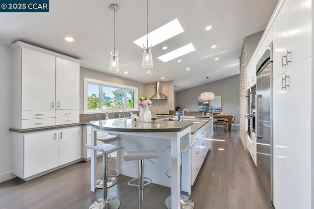 kitchen featuring hanging light fixtures, wall chimney exhaust hood, white cabinets, and a center island with sink