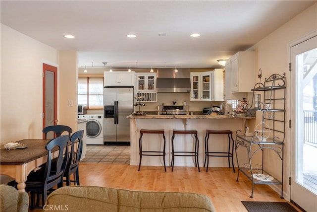 kitchen featuring white cabinetry, washer / dryer, kitchen peninsula, stainless steel refrigerator with ice dispenser, and wall chimney range hood