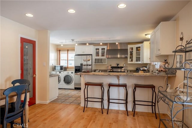 kitchen with wall chimney range hood, white cabinetry, stainless steel appliances, washing machine and clothes dryer, and kitchen peninsula