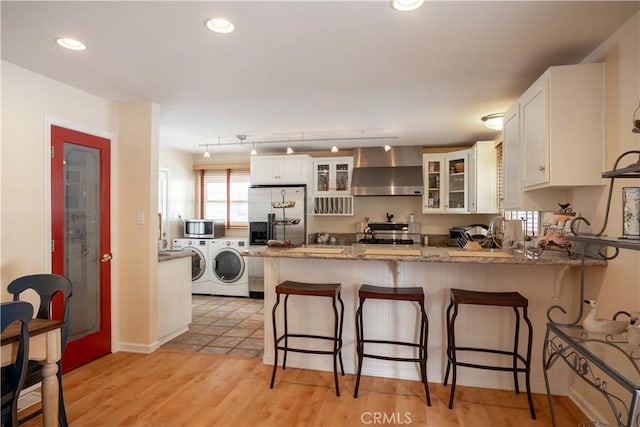 kitchen with washing machine and clothes dryer, wall chimney range hood, kitchen peninsula, stainless steel appliances, and white cabinets