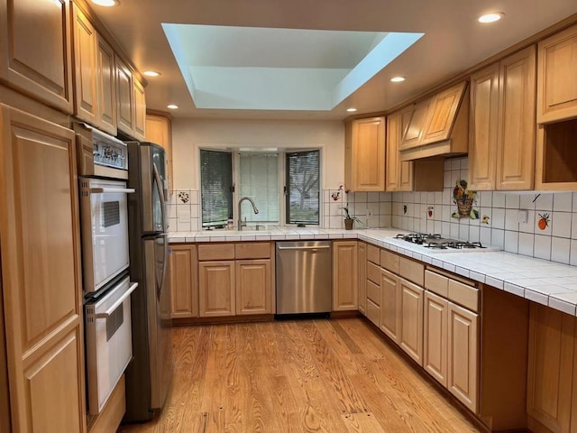 kitchen with premium range hood, appliances with stainless steel finishes, tile countertops, a raised ceiling, and light wood-type flooring