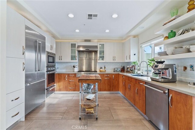 kitchen featuring range hood, white cabinetry, sink, backsplash, and built in appliances