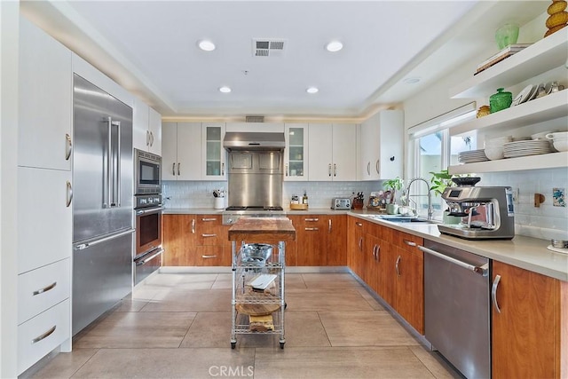 kitchen with glass insert cabinets, built in appliances, light countertops, white cabinetry, and a sink