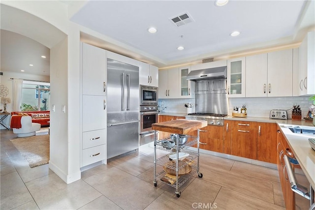 kitchen with range hood, white cabinets, decorative backsplash, built in appliances, and light tile patterned floors